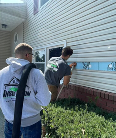 Two people injecting foam into the wall of a residential home from outside the house