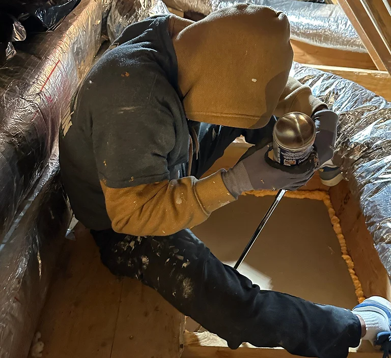 A worker in protective clothing kneels in an attic space, applying spray foam insulation around an opening between floor joists, with ductwork and insulation visible around them.