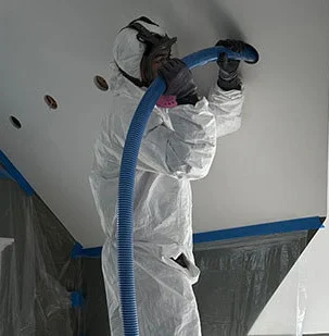 Worker in protective suit and respirator spraying insulation foam onto a ceiling in a prepared indoor space.